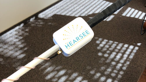 Close-up view of a white mobility cane prototype with a mounted Hearsee device, showing early-stage hardware development with a wrapped sensor wire.